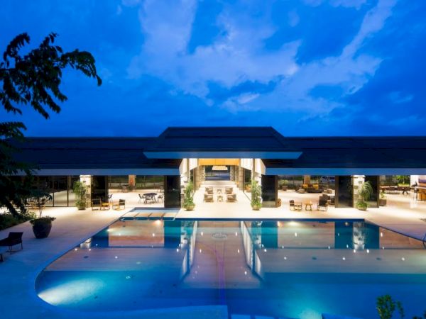 A luxurious, well-lit building with a spacious swimming pool at dusk, surrounded by trees and a clear sky.