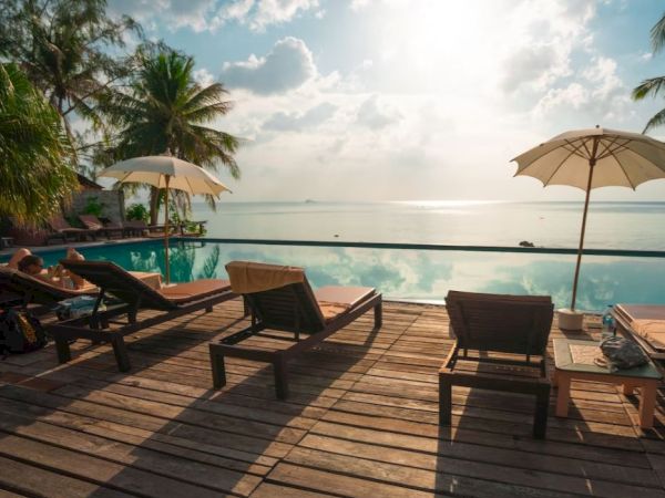 A tranquil poolside scene with lounge chairs, umbrellas, and palm trees overlooking a serene body of water under a partly cloudy sky.
