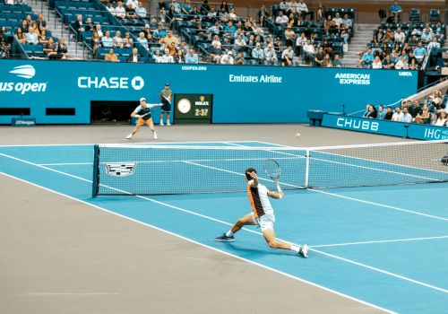 A tennis match at the US Open with two players on a blue court, surrounded by audience seating and sponsor logos visible.