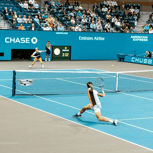 A tennis match at the US Open with two players on a blue court, surrounded by audience seating and sponsor logos visible.