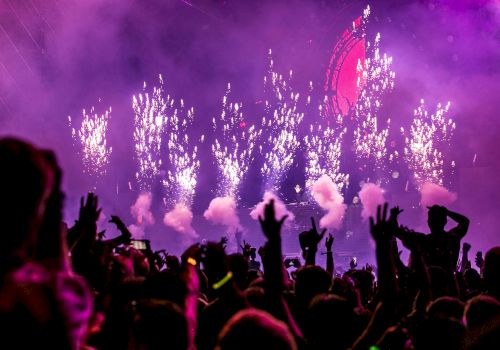 A vibrant concert scene with fireworks and cheering crowd, surrounded by pink and purple lights in an electrifying atmosphere.