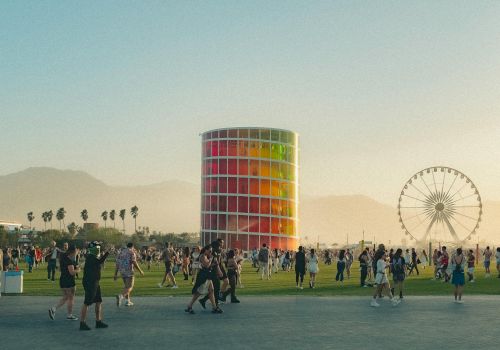 A vibrant outdoor scene with people gathered near a colorful structure and Ferris wheel, set against mountains and palm trees in the distance.