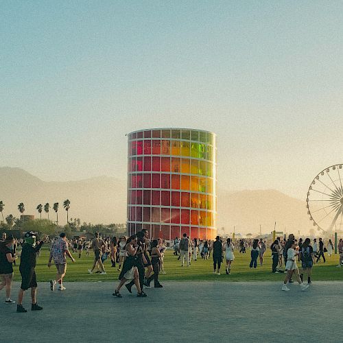 A vibrant outdoor scene with people gathered near a colorful structure and Ferris wheel, set against mountains and palm trees in the distance.