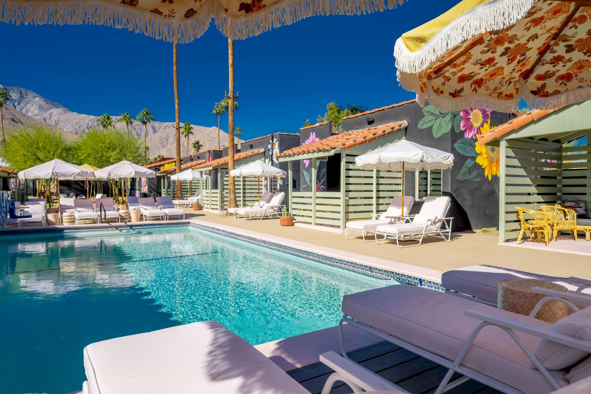 A sunny poolside area with lounge chairs, umbrellas, and colorful murals. Mountains and tall palm trees are visible in the background.