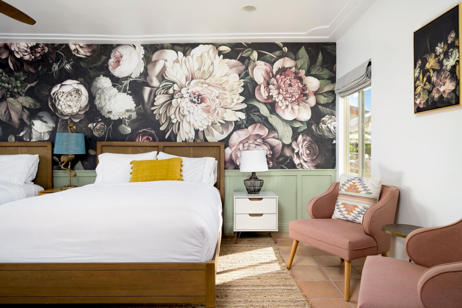 A bedroom with floral wallpaper, wooden bed, white bedding, pink chair, and a small table with a white lamp.