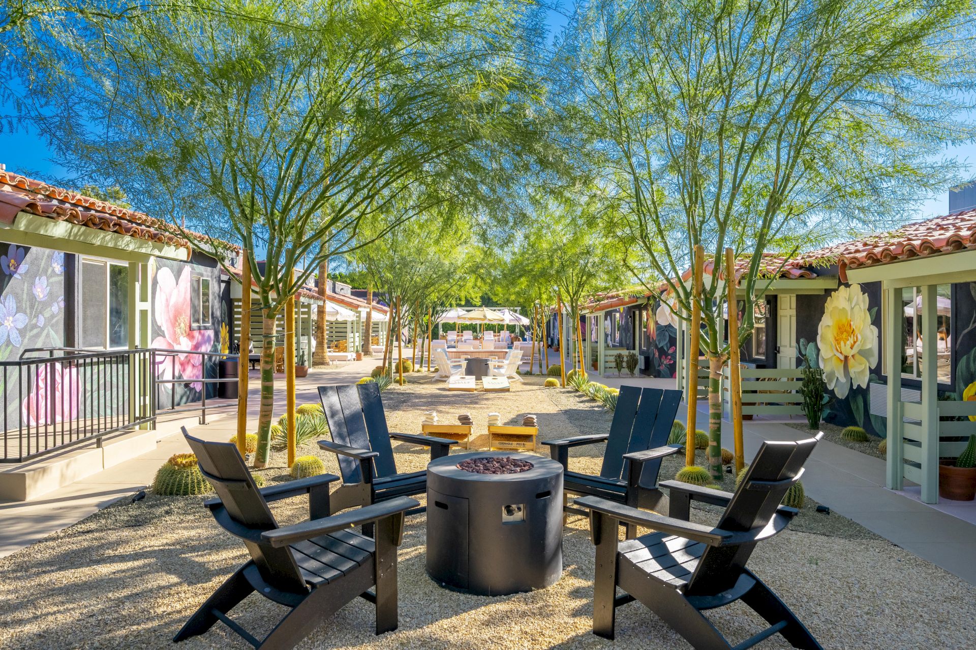Outdoor seating area with four chairs around a fire pit, surrounded by trees and small buildings with murals and decorative plants.