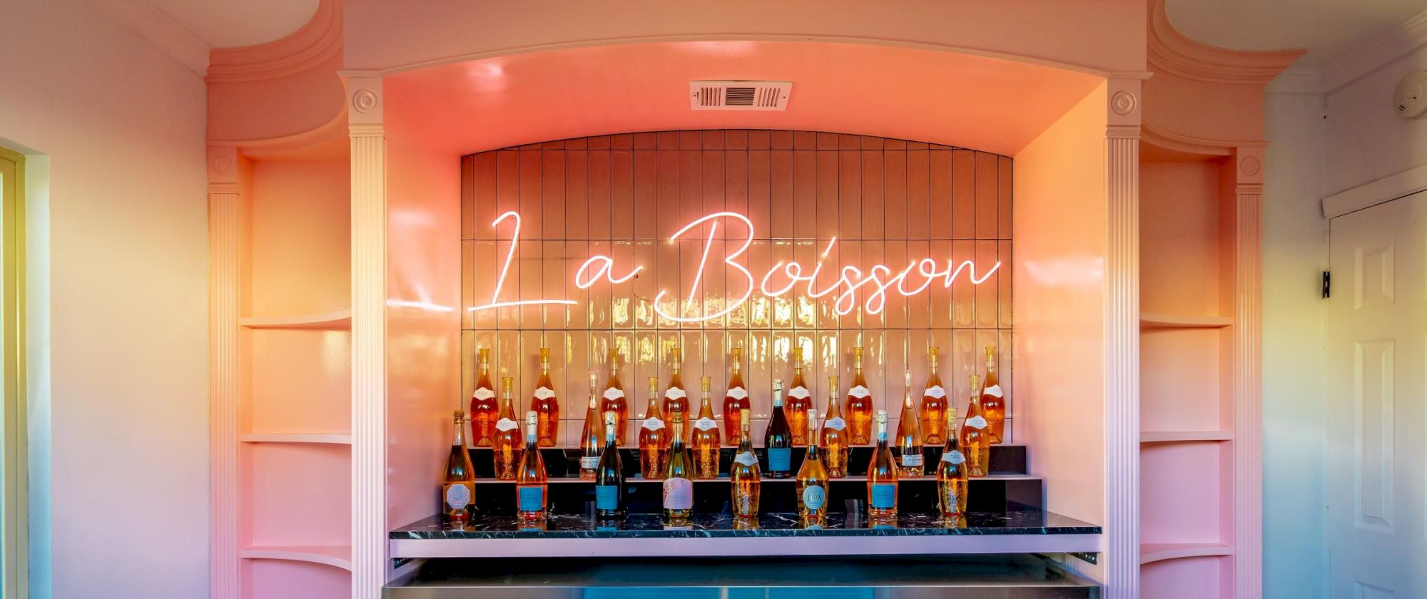 The image shows a pink-themed bar with bottles and a neon sign that reads "La Boisson" above them.