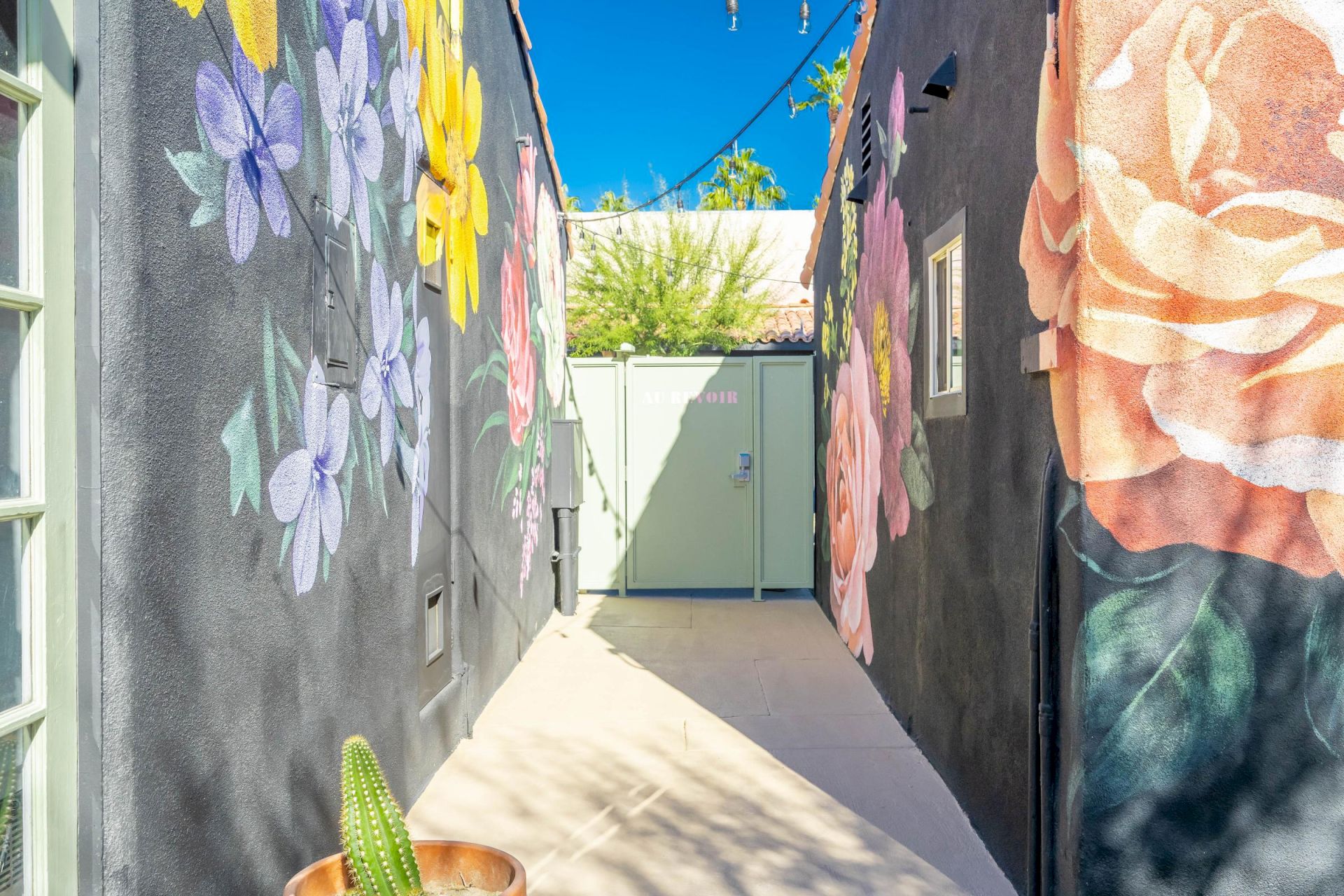 A narrow alleyway with colorful floral murals on both walls under a clear blue sky, leading to a closed gate.