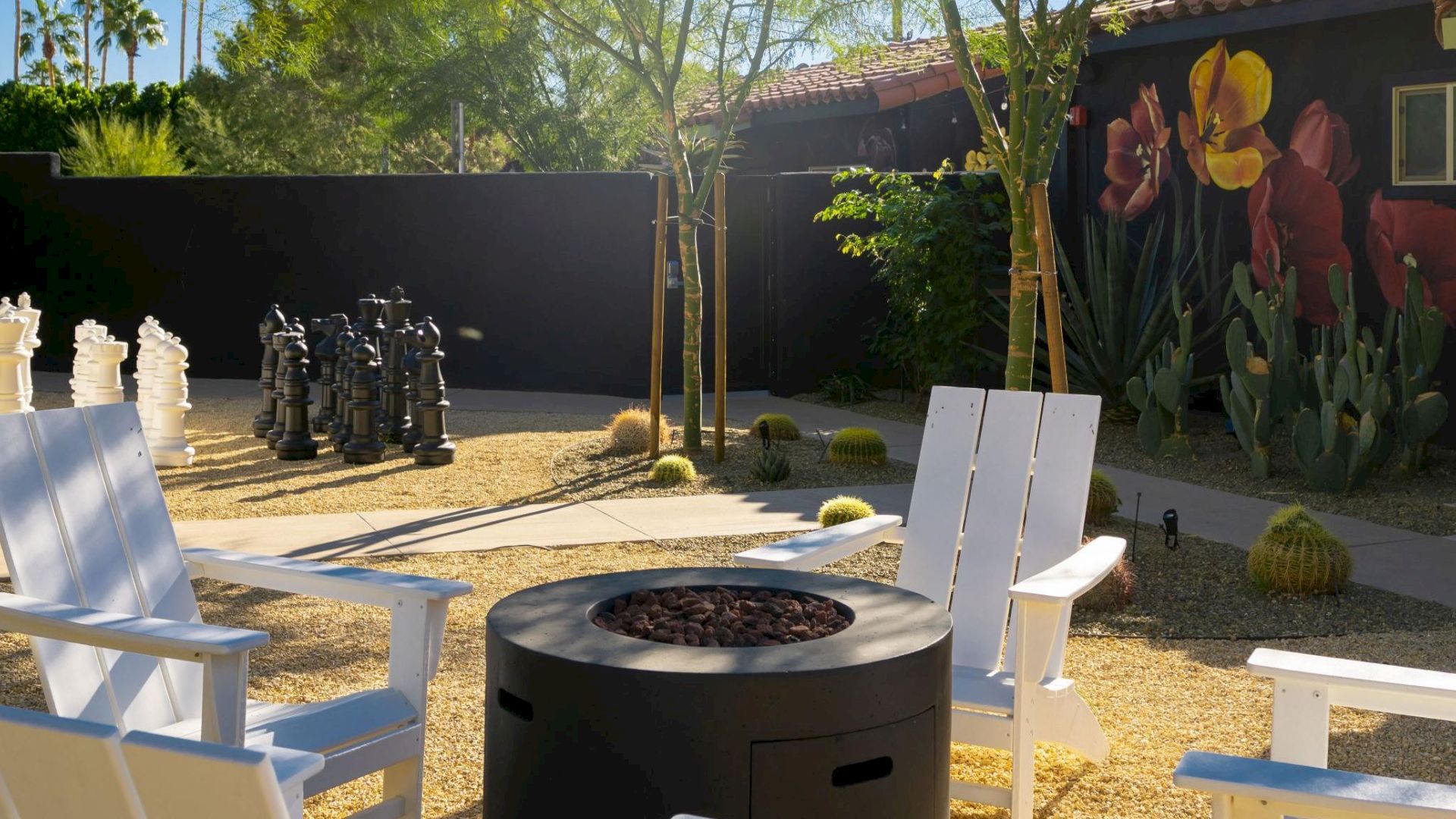 A sunny outdoor area with white chairs around a fire pit, large chess pieces, trees, and a mural with flowers on a building wall.