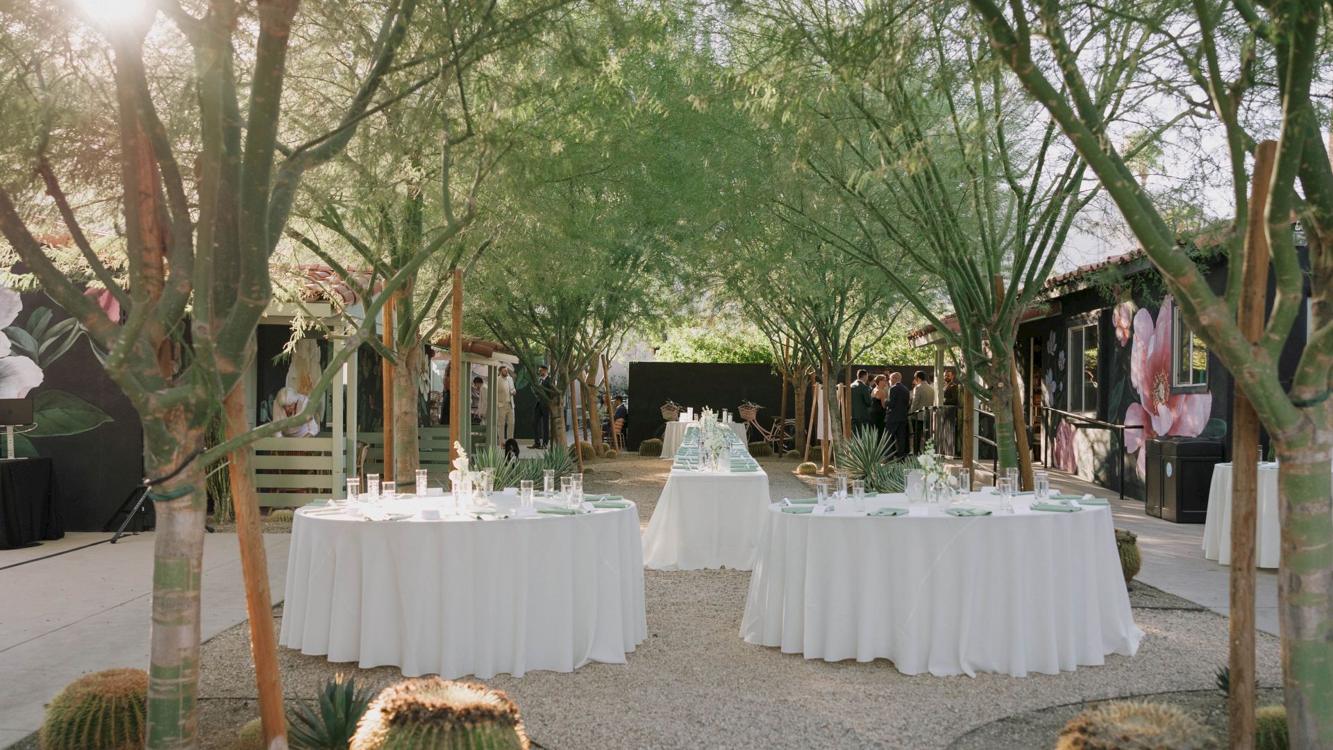 Outdoor event setup with round tables draped in white cloth amidst trees and greenery, surrounded by cacti and floral decorations.