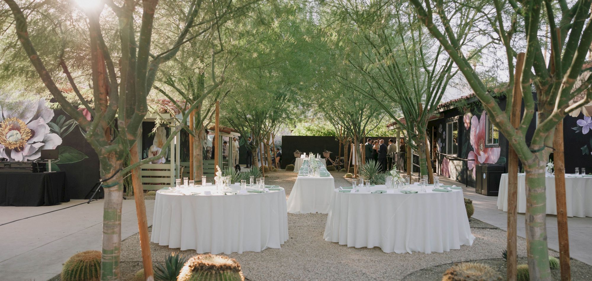 Outdoor event setup with round tables draped in white cloth amidst trees and greenery, surrounded by cacti and floral decorations.