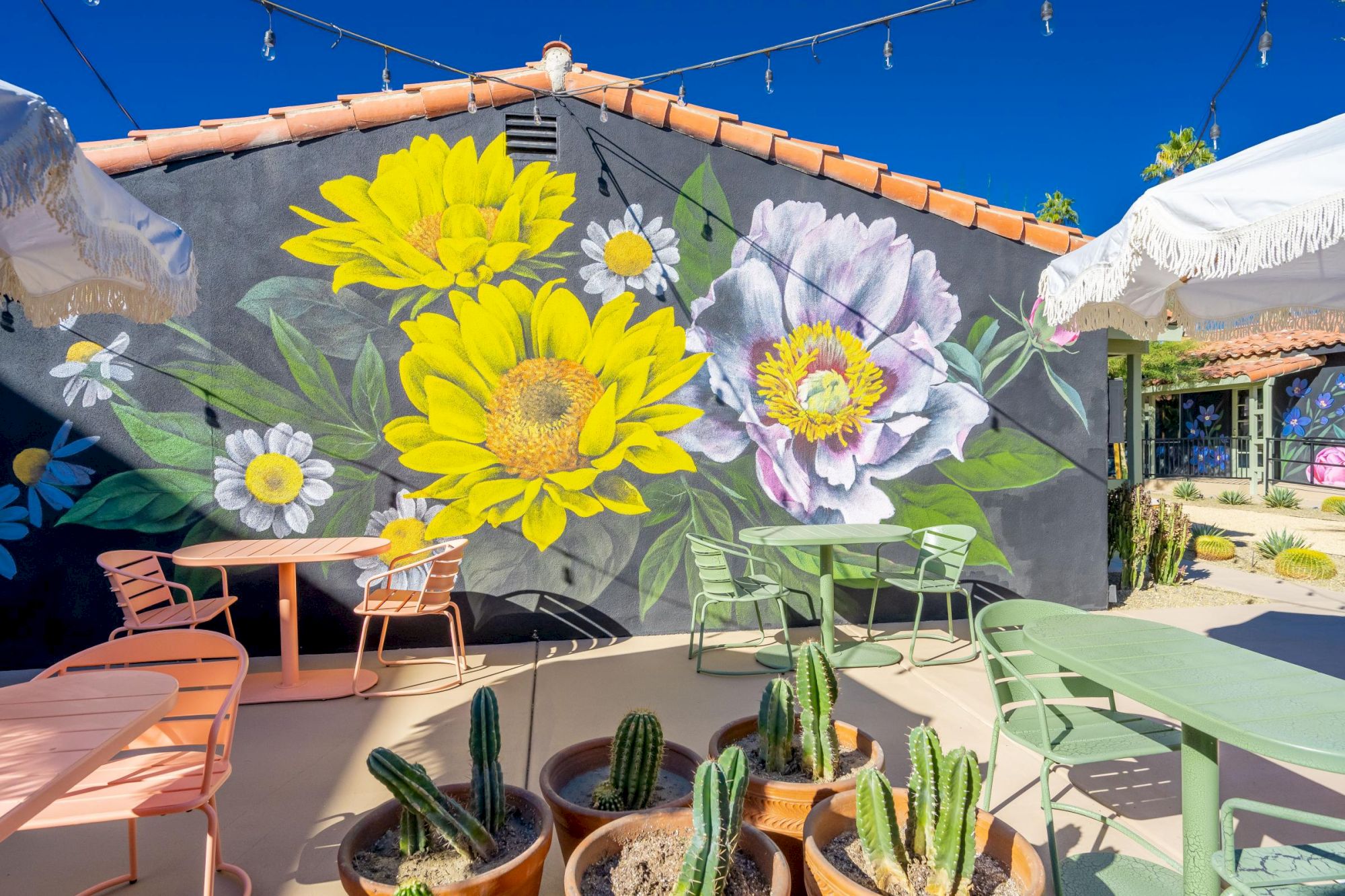 Outdoor seating area with colorful tables, chairs, potted cacti, and a large floral mural on the wall under string lights and umbrellas.