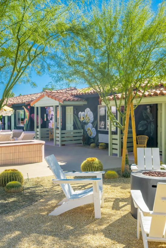 A sunny patio features white chairs, a fire pit, and desert plants. Colorful murals adorn the building walls under a clear blue sky.