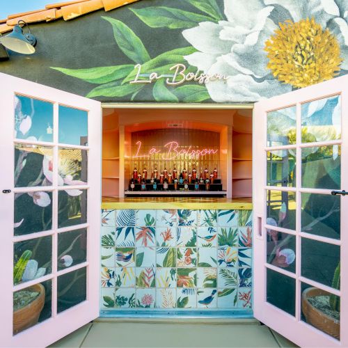 An outdoor bar with pink doors and floral wall art, displaying bottles inside under a neon sign. The facade is colorful and inviting.