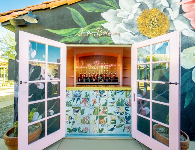 An outdoor bar with pink doors and floral wall art, displaying bottles inside under a neon sign. The facade is colorful and inviting.