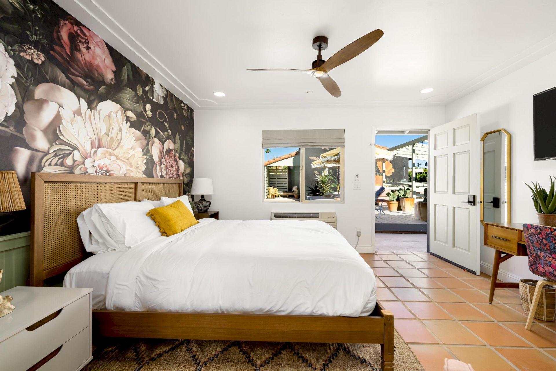 A cozy bedroom with a floral accent wall, bed with white linens and yellow pillow, ceiling fan, desk, and doorway leading outside.