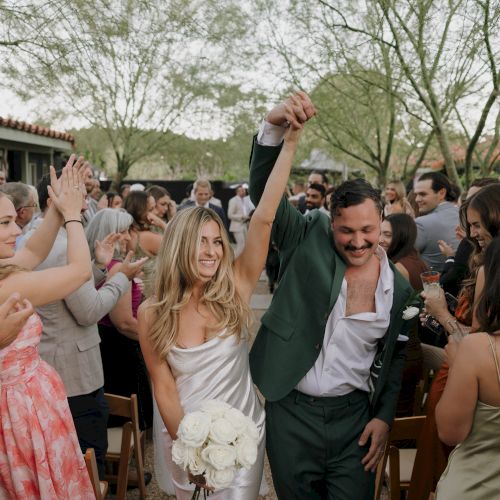 A couple celebrates at a wedding, surrounded by cheering guests. The bride holds flowers, and the scene is joyful and festive.