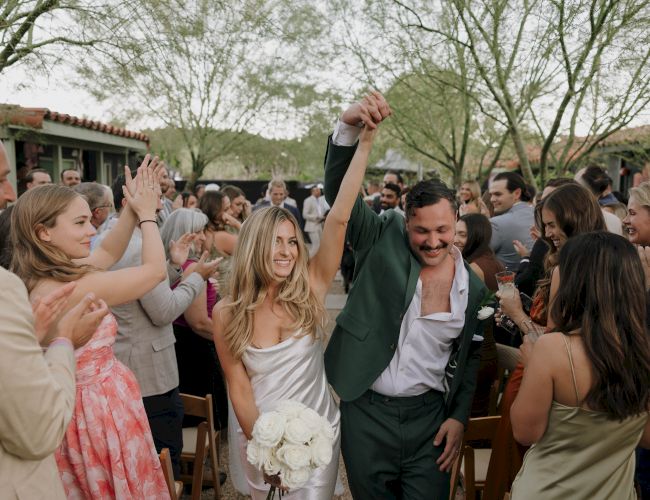 A couple celebrates at a wedding, surrounded by cheering guests. The bride holds flowers, and the scene is joyful and festive.