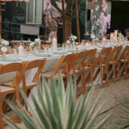 An outdoor dining setup with a long table, wooden chairs, candles, and flowers, surrounded by greenery in a garden setting.