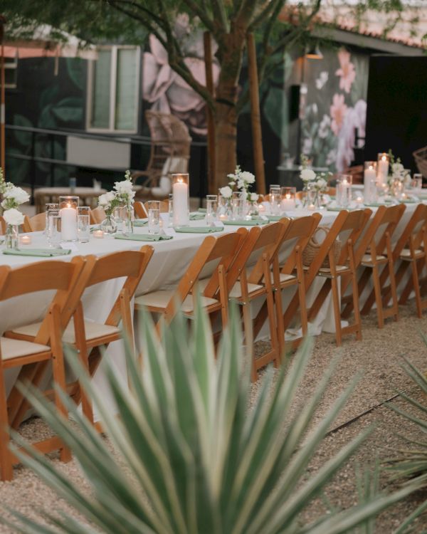 An outdoor dining setup with a long table, wooden chairs, candles, and flowers, surrounded by greenery in a garden setting.