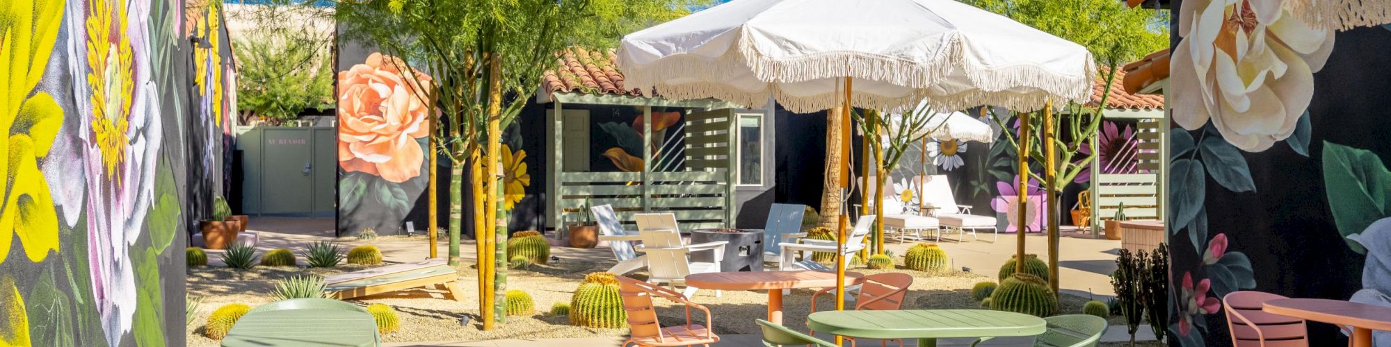 A vibrant outdoor patio with colorful chairs, tables, murals, potted cacti, and large white umbrellas under a sunny blue sky.