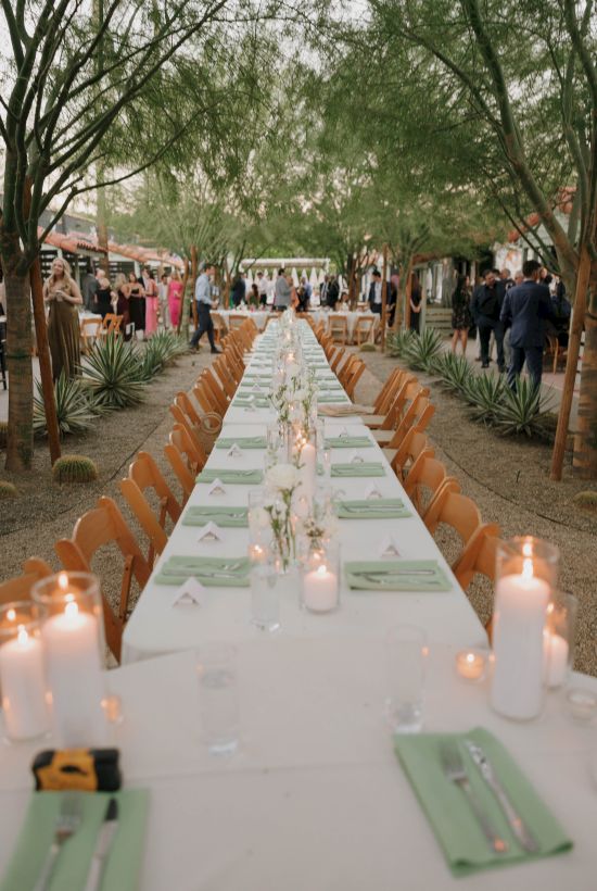 A long outdoor dining table is set with candles and green napkins, surrounded by trees and guests in the background.