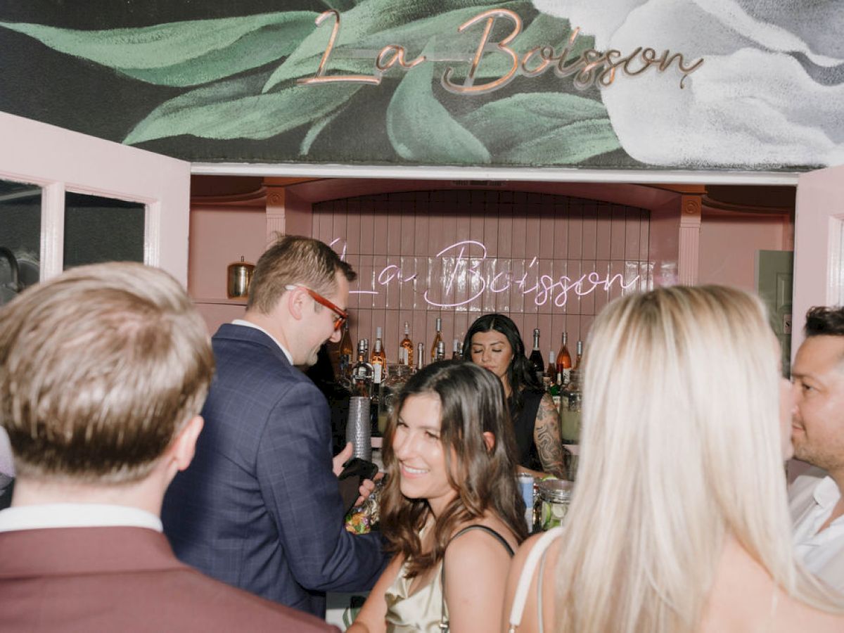 People gather in a bar setting with a floral sign above that reads "La Barsoon."