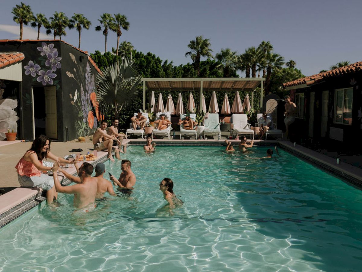 People are relaxing and swimming in a pool at a sunny resort with palm trees and lounge chairs in the background.