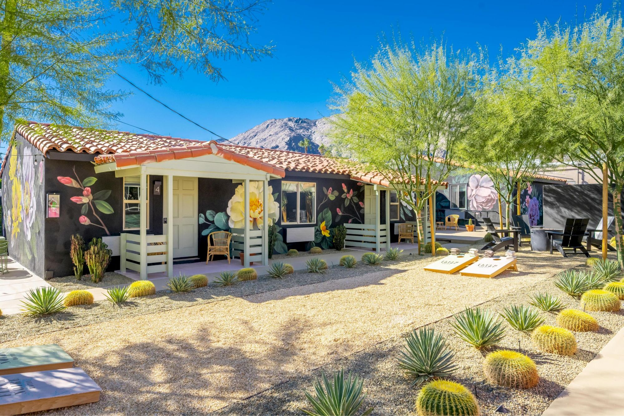 Small colorful cabins with murals are surrounded by desert landscaping, featuring cacti, trees, and a game setup in the courtyard.