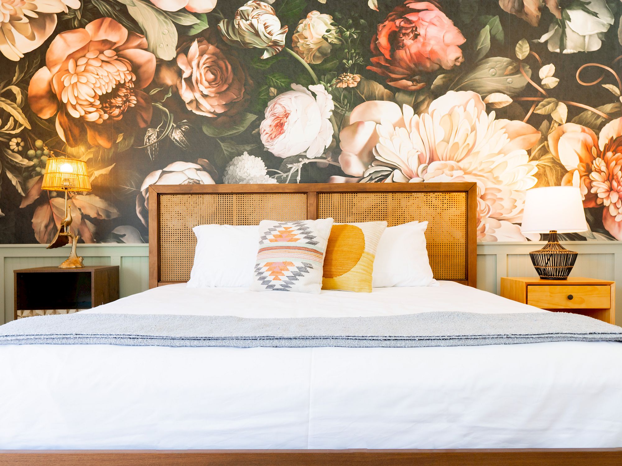 A bedroom features a floral mural wall, a wooden bed with white and patterned pillows, and bedside tables with lamps.