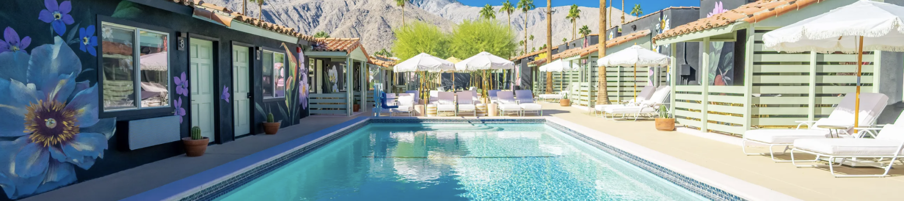 A sunny poolside with loungers, colorful parasols, and mural-adorned walls, set against a backdrop of mountains and clear blue skies.