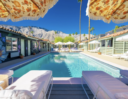 A sunny poolside with loungers, colorful parasols, and mural-adorned walls, set against a backdrop of mountains and clear blue skies.