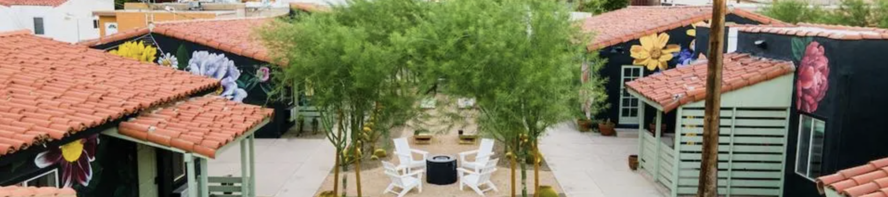 A courtyard with seating, surrounded by buildings with colorful murals, palm trees, and mountains in the background.