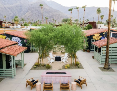 A courtyard with seating, surrounded by buildings with colorful murals, palm trees, and mountains in the background.