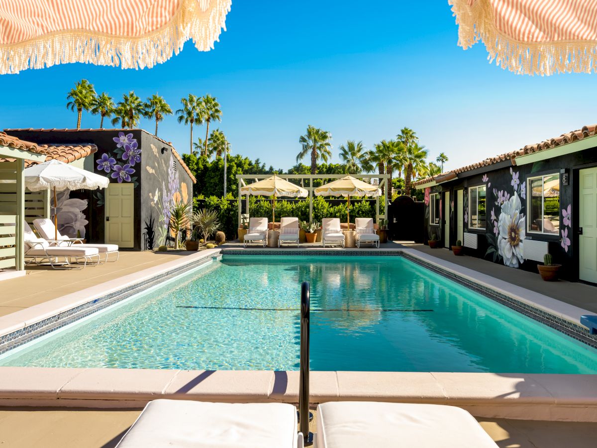 A luxurious outdoor pool area with sun loungers, umbrellas, and palm trees under a clear blue sky, creating a relaxing environment.