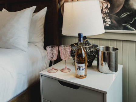 A bedside table with a lamp, two pink glasses, a bottle of wine, and an ice bucket.