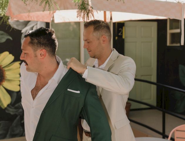 Two men are dressing in suits outdoors. One is helping the other with his jacket under a leafy tree and an umbrella.