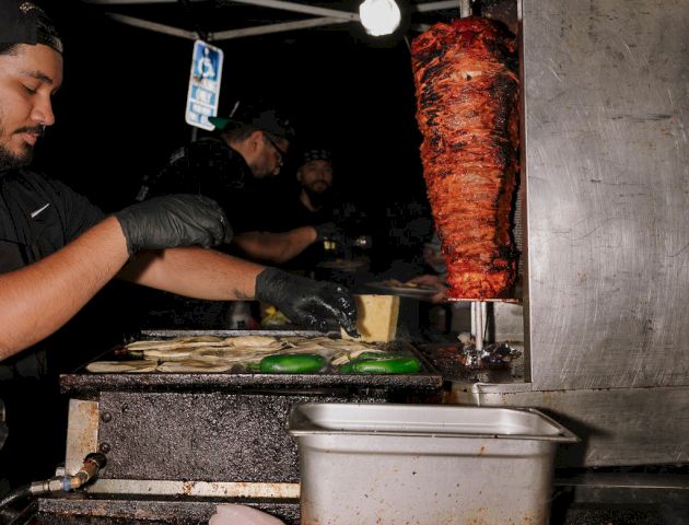 A street food setup with a spit of meat being cooked, raw fish fillets on a grill, and people preparing food in the background.