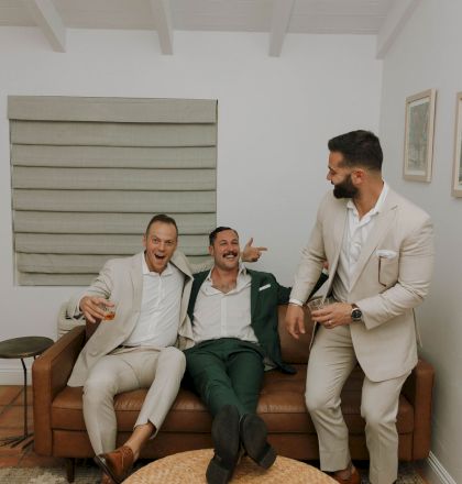 Three men in suits are laughing and holding drinks, sitting on a brown couch in a living room.