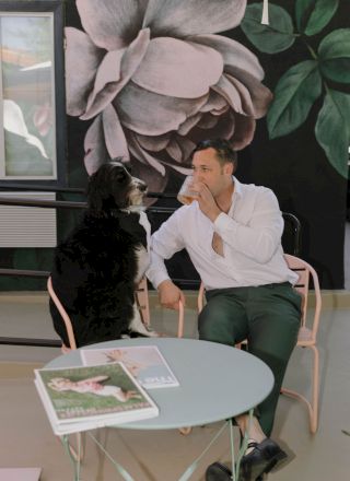 A man in a white shirt drinks from a glass while sitting next to a black and white dog in front of a floral mural.