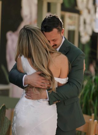 A couple is embracing, with the woman in a white dress and the man in a green suit, sharing a tender moment.