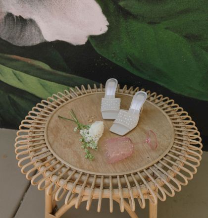 A wicker table with white shoes, a pink glass, white flowers, and gold rings, set against a green and pink mural background.