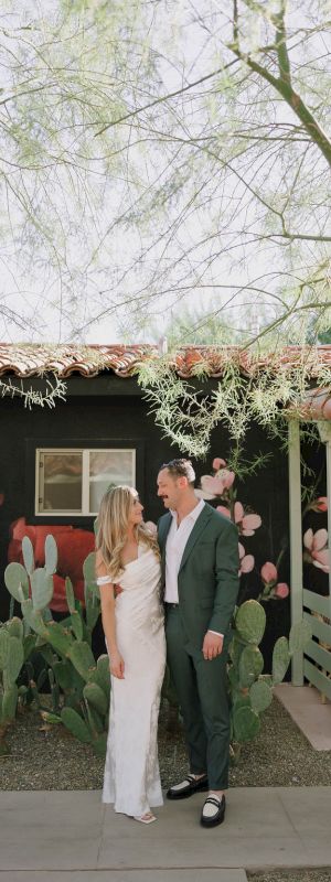 A couple stands in front of a mural with cacti, wearing formal attire, set against a house with terra-cotta roof tiles and greenery around.