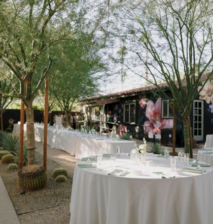 Outdoor gathering setup with round tables covered in white tablecloths, surrounded by trees and cactus plants, near a mural-adorned building.