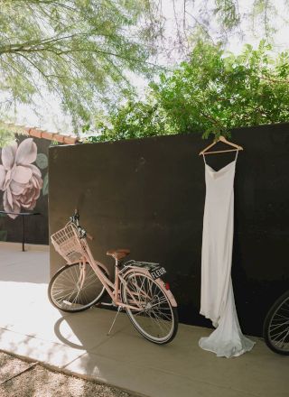 A pink bicycle with a basket leans against a black wall. A white dress hangs nearby. Greenery and a floral mural are visible in the background.