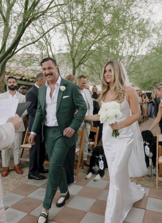 A couple walks down an outdoor aisle, the groom in a green suit and bride in a white gown, with guests and dogs present.