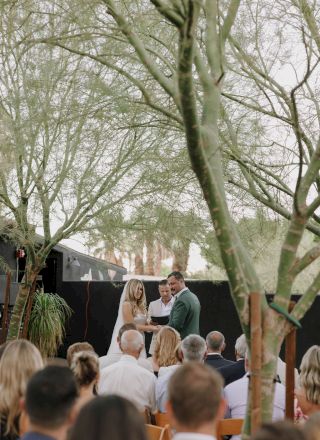 A couple getting married outdoors, surrounded by guests under green trees, with a minister officiating the ceremony.