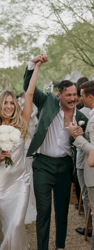 A joyous couple, one in a white dress with a bouquet, the other in a green suit, celebrating at an outdoor wedding ceremony.