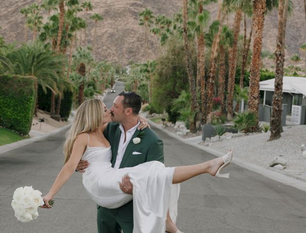 A couple is embracing on a palm-lined street, with the man carrying the woman in a white dress and holding a bouquet.