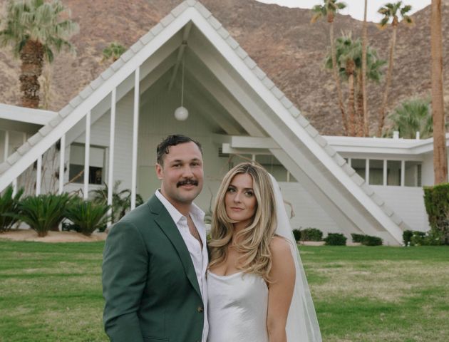 A couple in wedding attire stands in front of a white A-frame house with palm trees and mountains in the background.